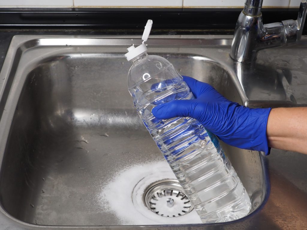 Sink drain cleaning process using baking soda and white vinegar. Close-up of a stainless steel kitchen sink and plastic bottle. Tips for home eco-cleaning - 1st Call Drain Clearance & Technical Services.